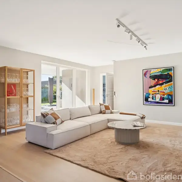 En beige sofa står på et stort tæppe i en stue. Sofaen vender mod et sofabord. Væggene har kunst og store vinduer slipper lys ind.