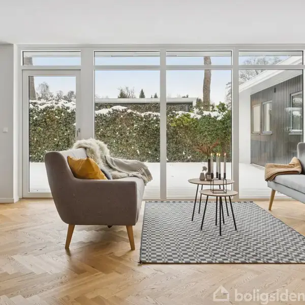 To sofaer står overfor hinanden med et sofabord imellem dem i en stue med trægulv. Store vinduer viser en snedækket have udenfor.