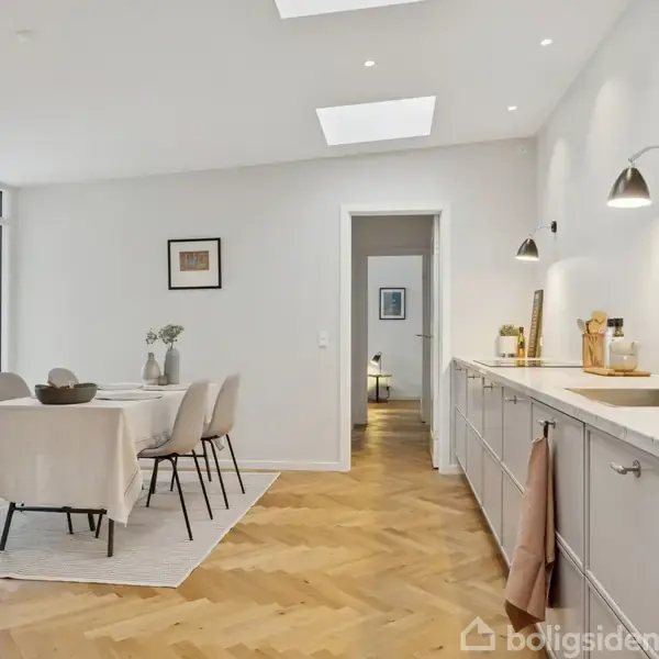 En spisestue med et bord dækket med dug og stole, ved siden af et langt køkkenbord med skabe, i et lyst rum med parketgulv og store vinduer mod haven.