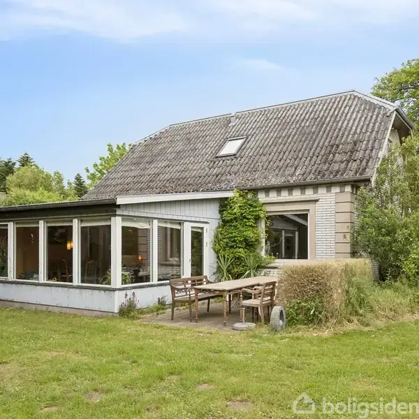 Huset har et skråt tag og store vinduer, er omgivet af grønne planter, og står på en græsplæne med havemøbler foran.