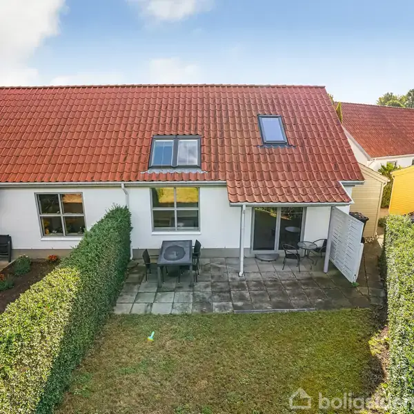 Et rækkehus med rødt tegltag og to skråvinduer. Foran huset er en flisebelagt terrasse med et bord og stole. Haven er omkranset af hække og et græsareal.