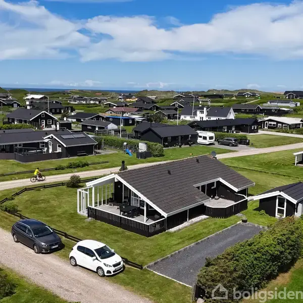 Et mørkt træhus med en terrasse står stille blandt andre huse i en kystnær sommerhusområde. To biler holder parkeret på grusvejen foran huset, mens en person cykler forbi.