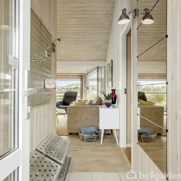 En lys gang med spejlvæg fører ind til en stue med sofaer og store vinduer, der giver udsigt til naturen udenfor. Skohylder og kurve står i gangen.