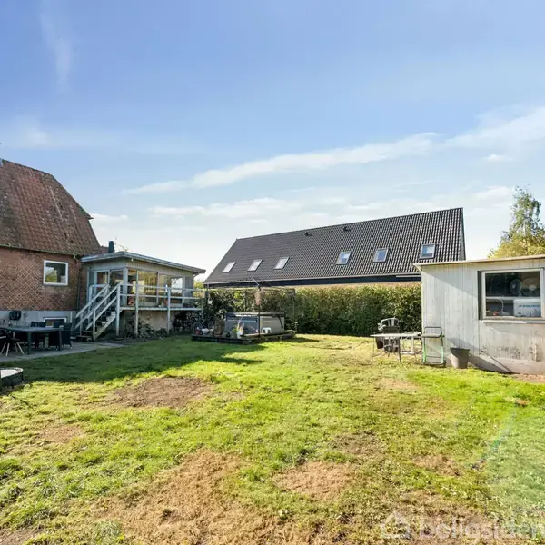 Et rødt murstenshus med en trappe til en baggård, der har en græsplæne og en skurbygning. Himlen er blå, og der er et sort tag i baggrunden.