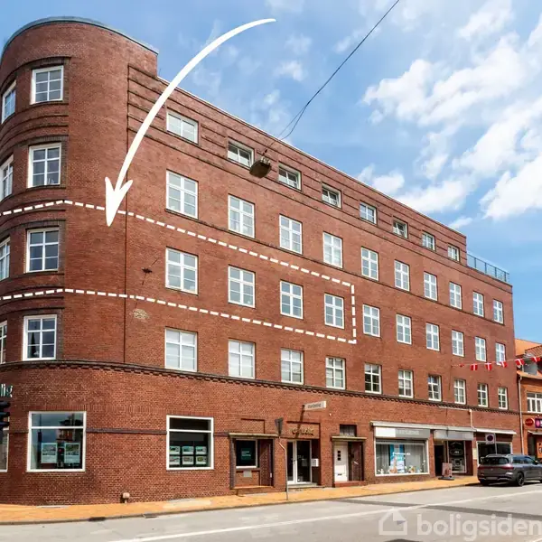 En rød murstensbygning med buet facade har en stor hvid pil og stiplet linje markeret på anden etage. Bygningen ligger på et gadehjørne med trafiklys og biler parkeret.