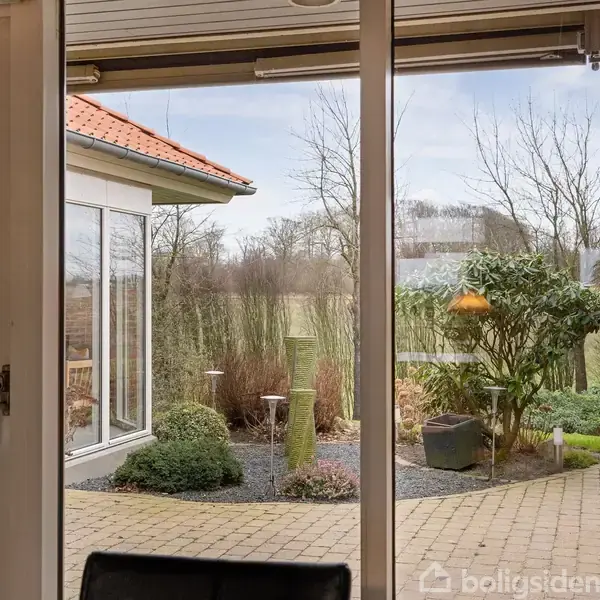 Glasdør viser en flisebelagt gårdhave med fuglehus og planter. Udsigten inkluderer mark og træer i baggrunden. Terrasseoverdækning og murstensvæg kan ses til venstre.