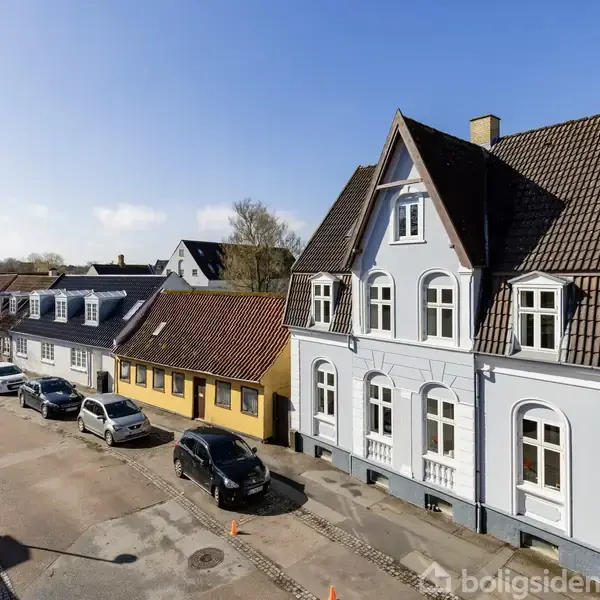 Flere farverige rækkehuse står side om side i en rolig gade. Biler er parkeret langs vejen under en klar, blå himmel med træer og bygninger i baggrunden.