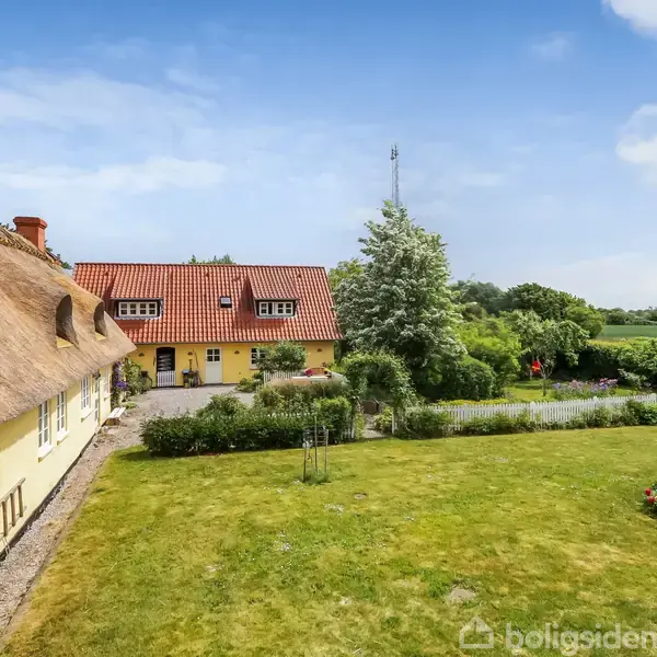 Stråtækt gul bygning med buede vinduer står stille i solbeskinnet have. Omkringliggende grønne planter og buske skaber en idyllisk landlig atmosfære under en klar blå himmel.
