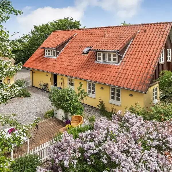 Et gult hus med rødt tag står blandt frodige buske og træer. Gårdspladsen er grusbelagt med cykler og en lille terrasse omgivet af en blomstrende have.