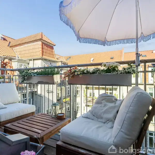 Terrasse med to komfortable loungestole og lille bord under en parasol. Terrassekasser med blomster hænger på gelænderet, omgivet af rødt tagstensbeklædte bygninger i et byområde.