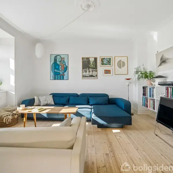Blå sofa står i en stue med trægulv, omgivet af kunst på væggene. Et sofabord står foran sofaen, og der er en fladskærms-TV til højre. Store vinduer slipper lys ind.