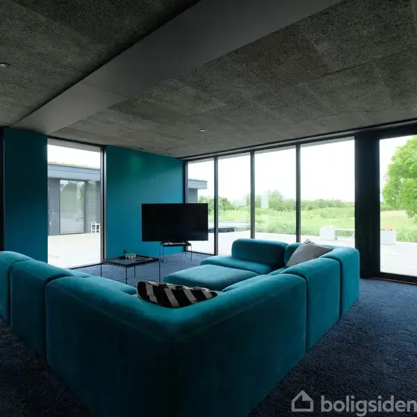 En stor, blå sofa står i midten af en stue og vender mod et fjernsyn. Stuen har store vinduer, der giver udsigt til en terrasse og grønne omgivelser.