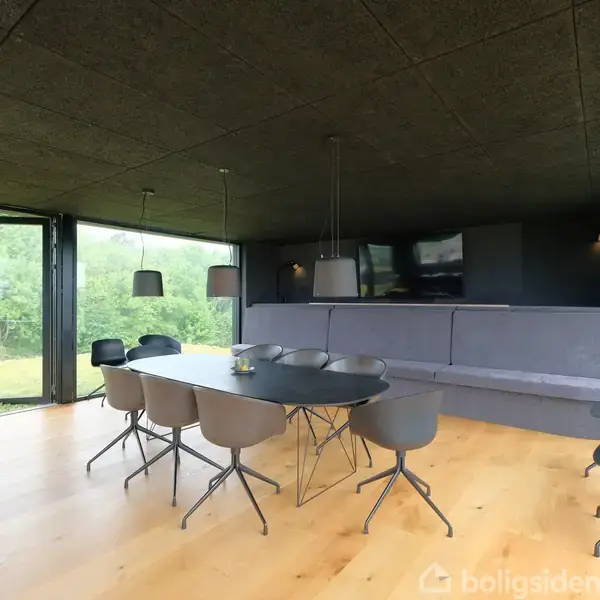 Spisebord med seks stole står i et moderne, lyst rum med store vinduer, der giver udsigt til frodig natur udenfor. En lang, grå sofa står ved den ene væg.