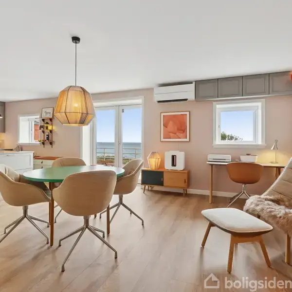 Et moderne køkken-alrum med spisebord, otte stole og pendellamper er centralt placeret. Store vinduer giver udsigt. Rummet er lyst og indrettet med minimalistiske møbler.