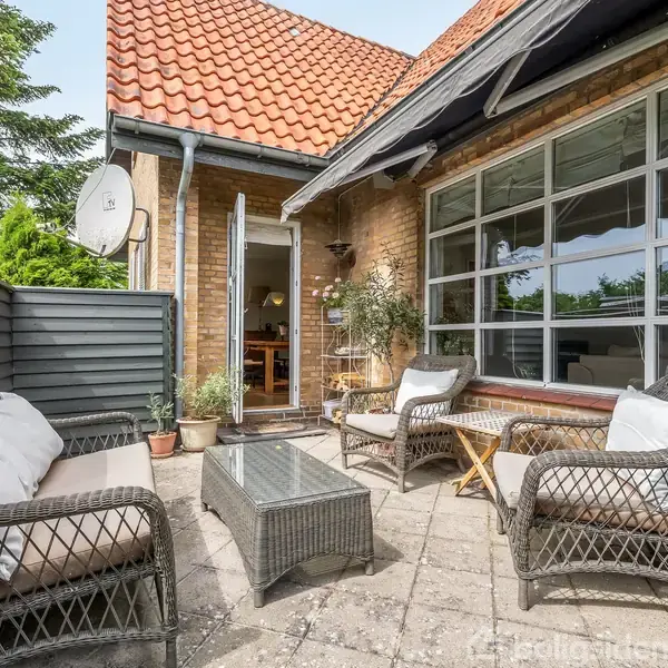 Terrassemøbler står på flisebelagt område med ratansofaer og bord, dekoreret med puder, foran en murstensbygning med store vinduer og åben dør. Omkranset af planter og træhegn.