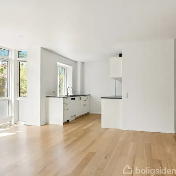Et lyst, tomt rum med trægulv, to vinduer til højre, der slipper sollys ind, og en radiator under det ene vindue. Væggene er hvide, og rummet er minimalistisk.