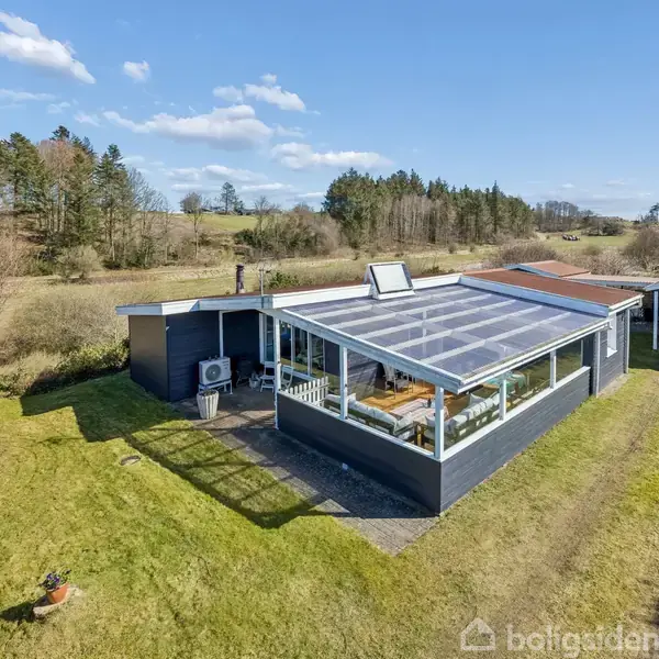 Et sommerhus med et stort glasoverdækket terrasseområde står midt i en grøn have. Bygningen er omgivet af åbent landskab med spredte træer i baggrunden.