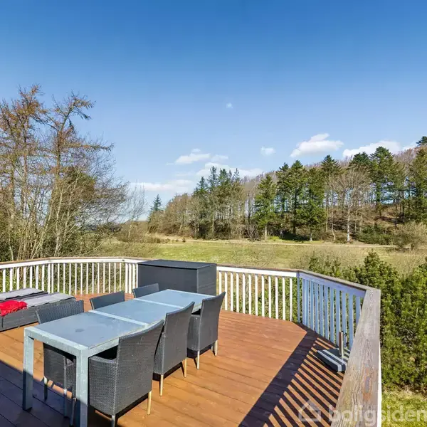 Terrasse med møbler, herunder sofa og bord, placeret på trædæk. Solseng ved siden af sofaen. Baggrund viser grønne træer og en klar blå himmel i et skovområde.