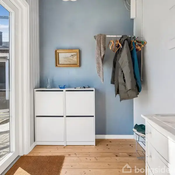 En entré med lyseblå vægge, et skoskab og hængende jakker. Et maleri hænger over skoskabet, og der er udsigt til en have gennem en stor glasdør. Gulvet er i lyst træ.
