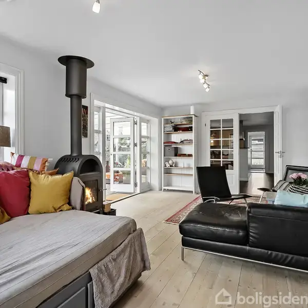 Brændeovn står i stuen og opvarmer rummet, omgivet af en beige sofa med farverige puder, sorte lænestole og en stor bogreol. Glasskydedøre fører ud til terrassen.