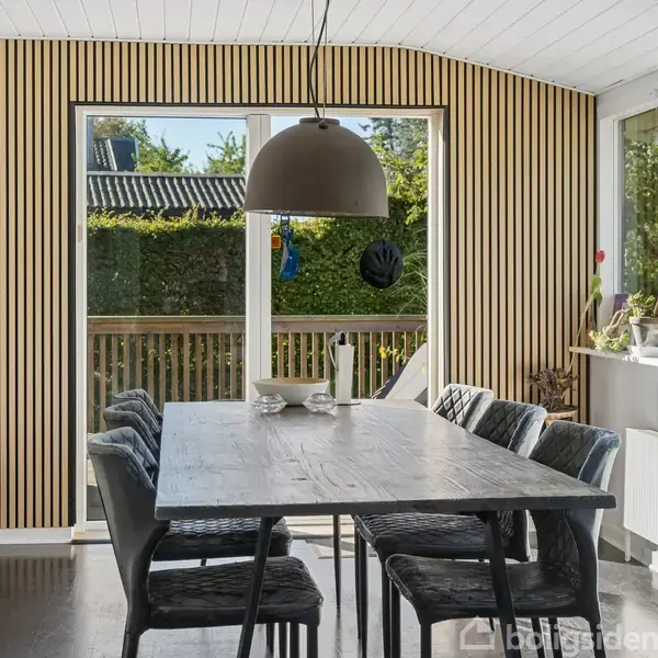 Et spisebord står omgivet af stole i et lyst rum. En stor lampe hænger over bordet. Rummet har trævægge og store vinduer, der vender ud mod en grøn hæk.