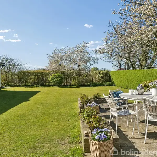 En udendørs terrasse med havemøbler er omgivet af frodigt grønt græs og blomstrende træer under en klar blå himmel. Der er blomster i potter på og omkring bordet.