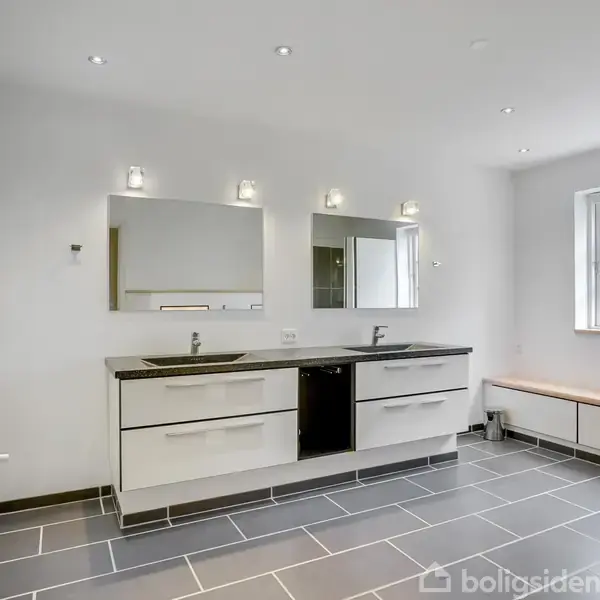 Badeværelse med dobbeltvask i en sort bordplade, to spejle og hvide skabe under. Der er flisegulv, en bænk ved vinduet og hvidmalede vægge.