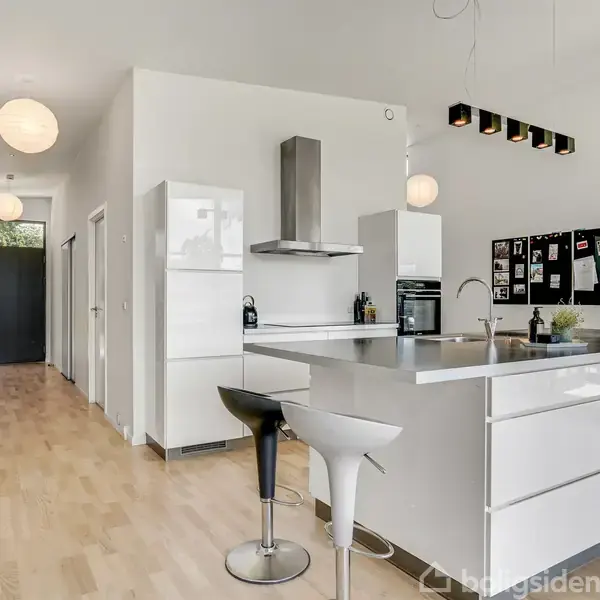 Køkken med hvidt, moderne interiør indeholder en ø med to barstole. Omgivet af træplankegulv, vægdekorationer, og opslagstavler. Store runde lamper hænger fra loftet.