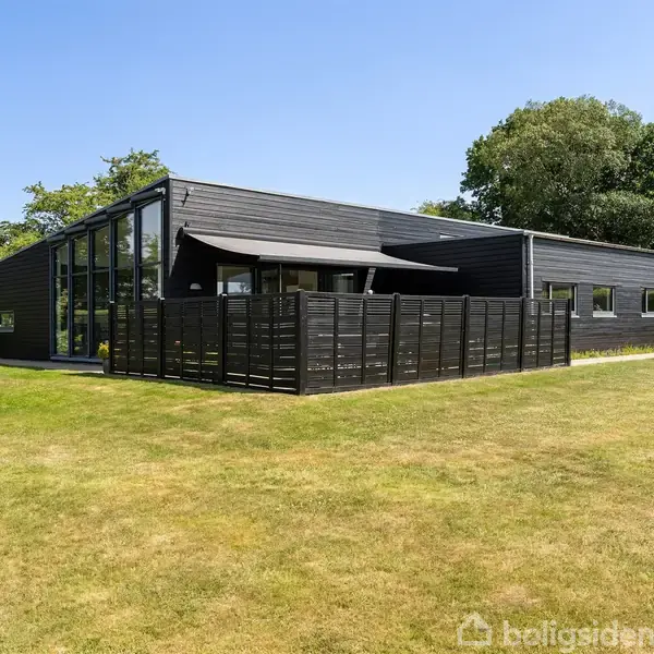 En moderne, sort træbygning står i en solrig have omkranset af træer. En indhegnet terrasse er på bygningens venstre side, mens græsplænen strækker sig rundt omkring.