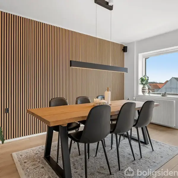 En spisestue med et langt træbord og seks sorte stole, omgivet af en moderne væg med lodrette paneler. En stor lampe hænger over bordet, og der er adgang til en balkon med udsigt over hustage. En grøn plante står i hjørnet.