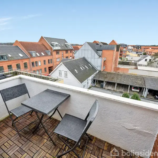 En lille altan med et sort cafésæt består af et bord og to stole, og den tilbyder udsigt over en by med røde murstensbygninger under en klar blå himmel.