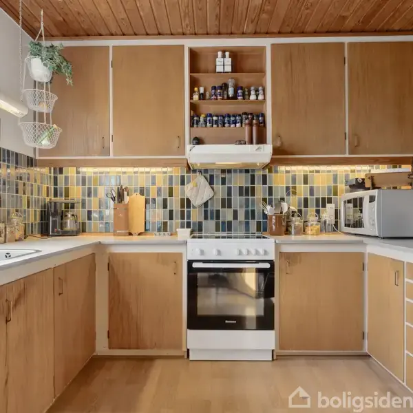 Et kompakt køkken med lyse træskabe fyldt med krydderiglas over komfuret. Der er en vask ved vinduet til venstre og en mikrobølgeovn til højre. Fliser i blå nuancer på væggen.