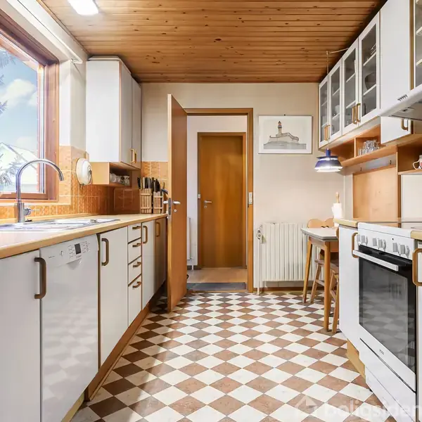 Køkken med hvide skabe og brun bordplade, hvor en opvaskemaskine er placeret ved siden af en vask. Skakternet gulv og træloft giver et retro look.