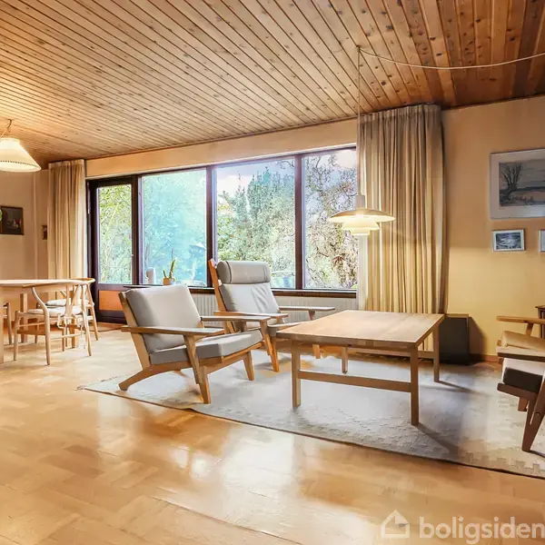 Stue med træmøbler, herunder to lænestole og en sofa, omkring et sofabord på tæppe. Store vinduer og træloft skaber lys og varm atmosfære. Bord og stole ses i baggrunden.