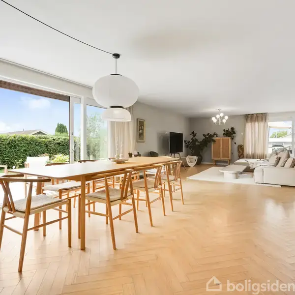 Spisebord med stole står i en rummelig og lys stue med trægulv. Store vinduer slipper naturligt lys ind, og en sofa er placeret i baggrunden ved siden af planter.
