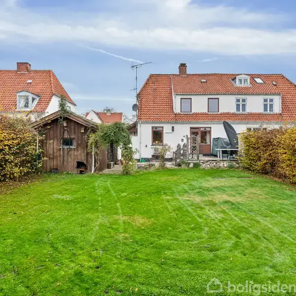 Hus med rødt tegltag, placeret bag en velplejet græsplæne, flankeret af tætklippede hække og et lille træskur til venstre. Blå himmel og enkelte skyer i baggrunden.