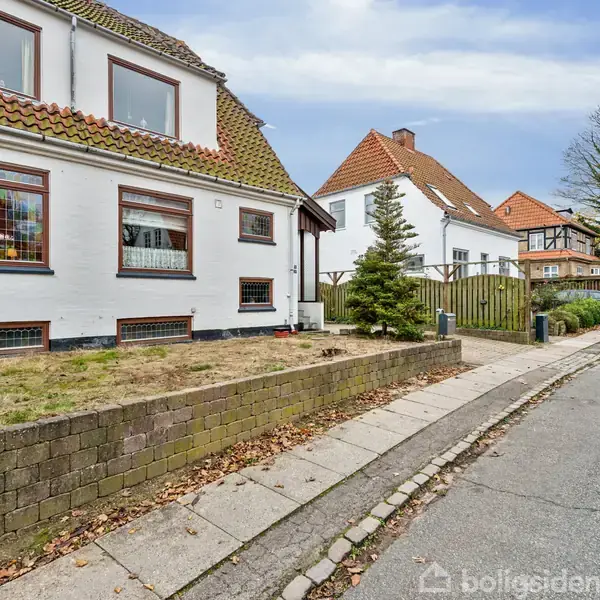 En hvidmalet murstenshus med brunt tegltag står ved en stille vej. Naboer omfatter lignende villaer, og fortovet er omgivet af små buske og græsplæner.