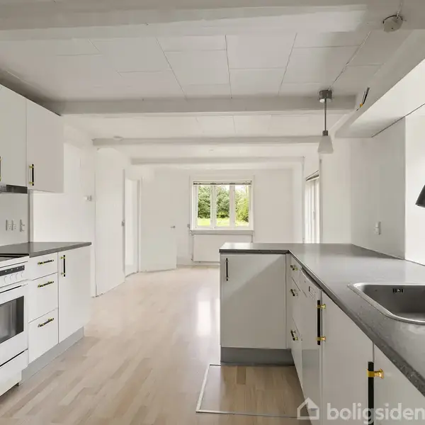 Et lyst, moderne køkken med hvide skabe og grå bordplader. En vask med hane er installeret i bordpladen. Store vinduer giver udsigt til grønne omgivelser udenfor.