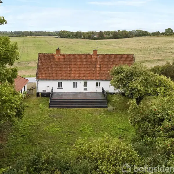 Hus med rødt tegltag og hvid facade står stille i naturskønt landskab med marker i baggrunden og omgivet af grønne træer.