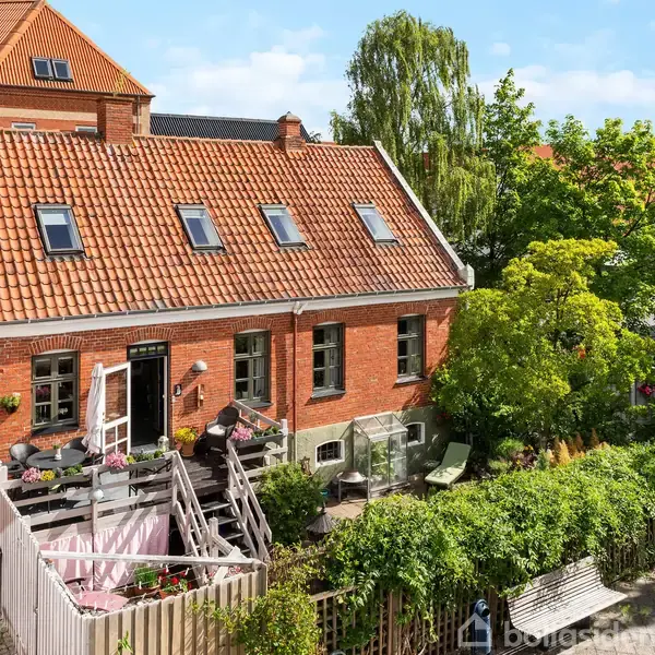 Rødt murstenshus med rødt tegltag, placeret i en frodig have med træer og buske. Ved siden af huset er der en terrasse med møbler. Bygninger ses i baggrunden.