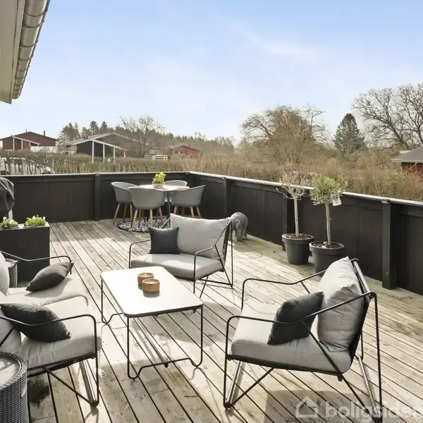Terrasse med træmøbler og puder står på en trædæk med sort rækværk. En bordgruppe og planter i potter skaber en indbydende stemning med udsigt over grønne omgivelser og huse i baggrunden.