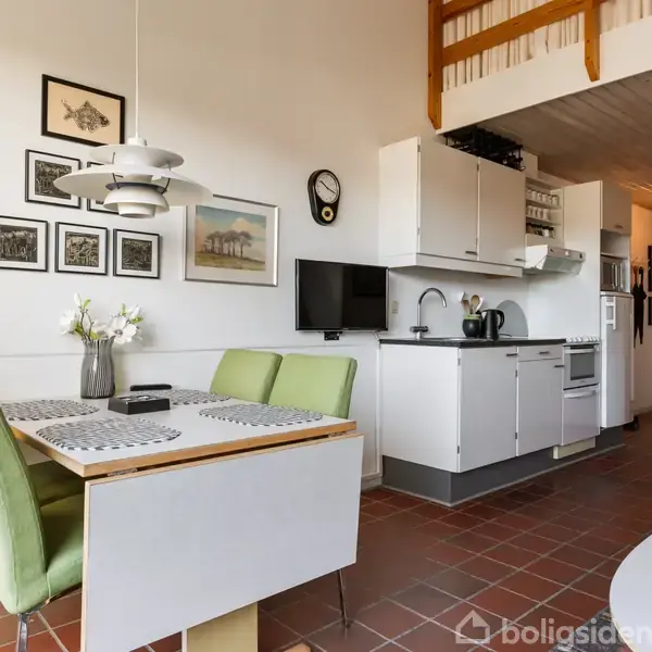 Et moderne køkken-alrum med et spisebord dækket med dækkeservietter og blomstervase. Grønne stole står ved bordet. Væggene har indrammede billeder, og der er et vægur.