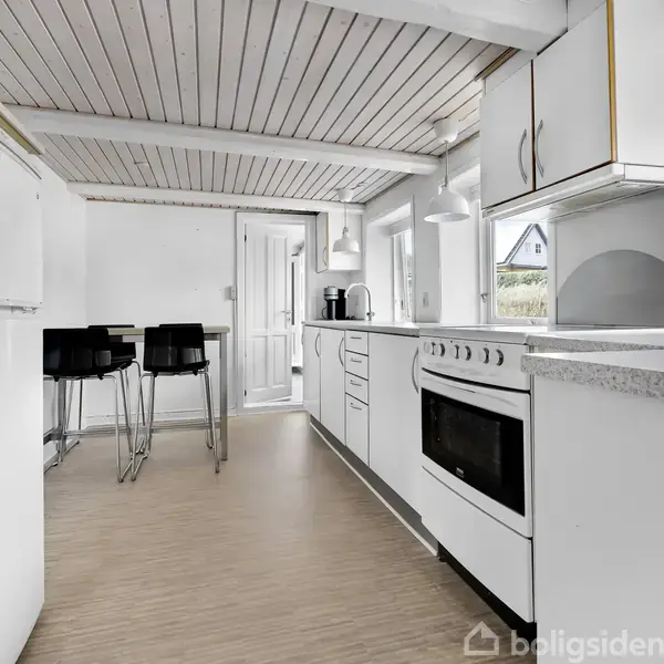 Et moderne køkken med hvide apparater og skabe. En ovn er centreret, og en spiseplads med høje stole er til venstre. Sollys kommer ind gennem vinduerne.