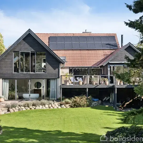 Et stort, moderne hus står omgivet af en velplejet have. Huset har store vinduespartier og solpaneler på taget. En terrasse med møbler ses på første etage.
