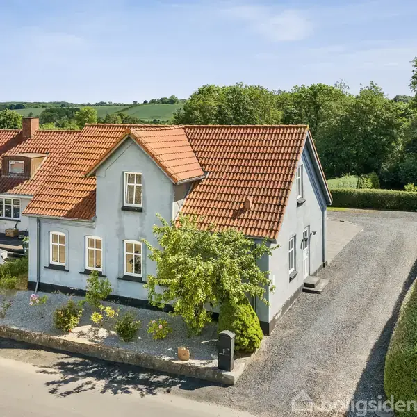 Et hus med rødt tegltag står på en grusbelagt grund, omgivet af frodig vegetation. Ved siden af huset holder en hvid bil, og til venstre ses et andet hus.