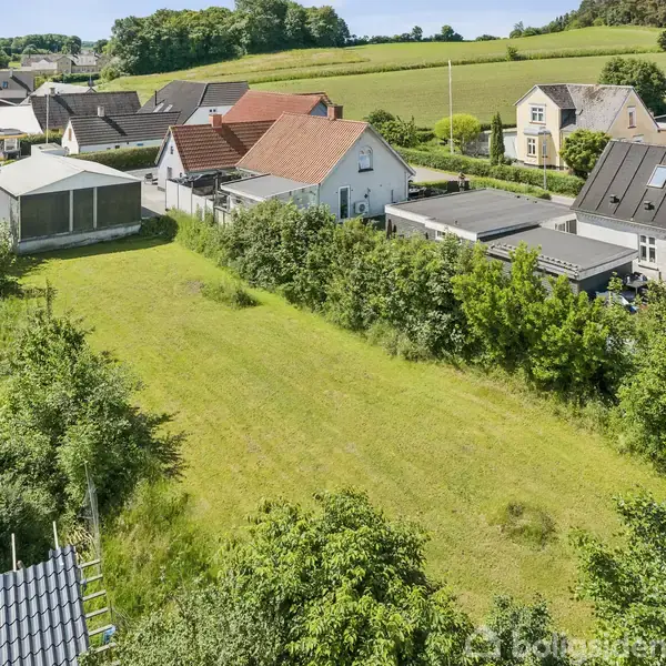 Grønt areal omkranset af huse, træer og buske. Omkringliggende bygninger har skrå og røde tage. Bagved ses marker og skov. Sollys giver et lyst og frodigt udtryk.