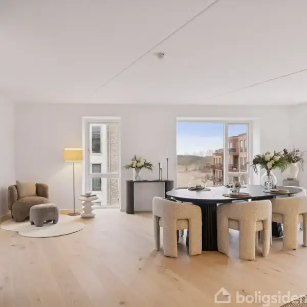 Stue med rundt spisebord og seks stole, blomsterdekoration på bordet. Lænestol og lampe i hjørnet. Store vinduer med udsigt til bygninger.
