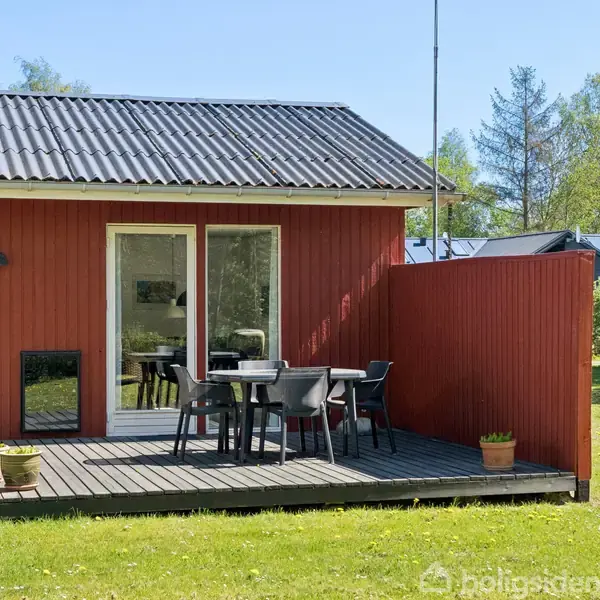 Rødmalet hus med en lille terrasse, hvor bord og fire stole står. Omgivet af en grøn græsplæne og træer i baggrunden. En potteplante står ved siden af terrassen.