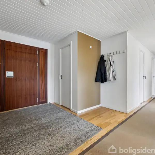 En rummelig entre med en stor dør til venstre, dekorative planter på en skænk og jakker hængende på kroge. Lyse trægulve og hvid loftbeklædning skaber en lys atmosfære.