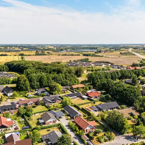 Boliger med farverige tage ligger spredt i en grøn forstad, omgivet af træer og åbne marker. Horizonterne strækker sig over et fladt landskab under en klar blå himmel.